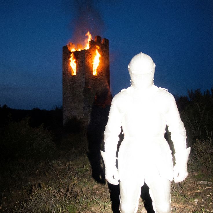 A knight in white armor stands at night before a burning stone tower.